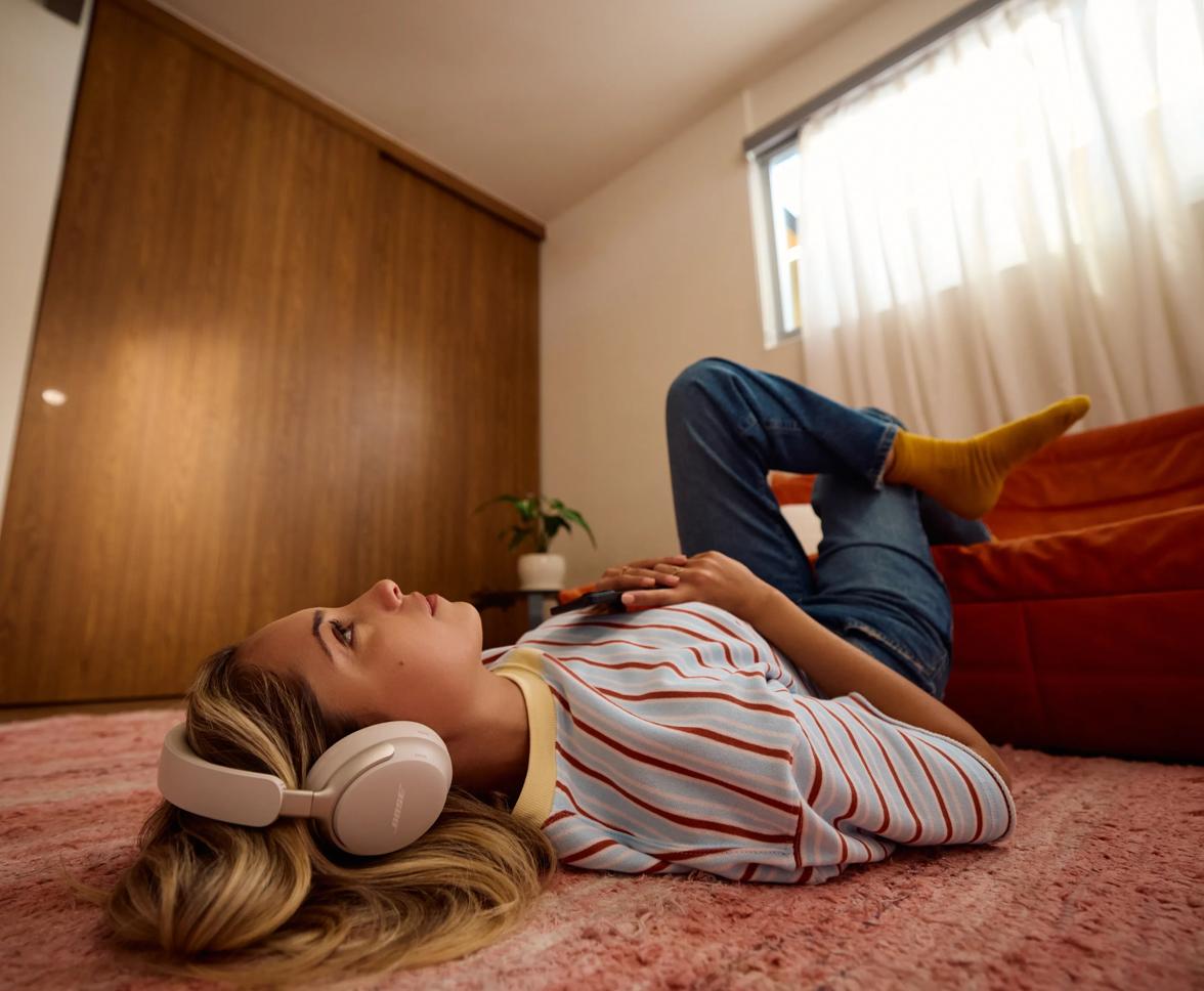A woman wearing QuietComfort Ultra Headphones in White Smoke sprawled out on the floor of a living room listening to music.
